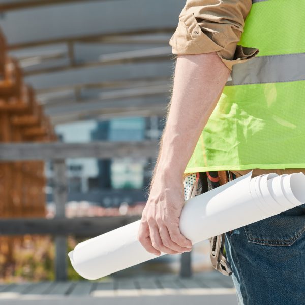 Rear view of unrecognizable construction man in green vest standing against unfinished bridge and holding blueprint