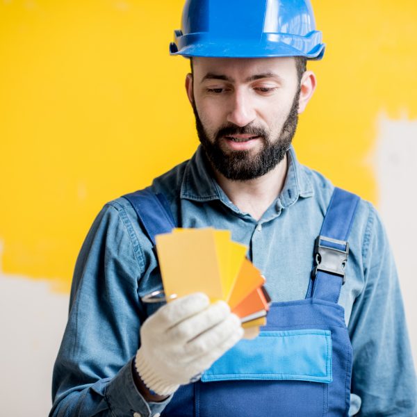 Painter in blue workwear holding color swatches on the yellow wall background indoors